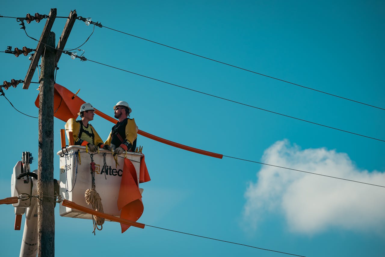 Two electricians in safety gear working on a utility pole with a clear blue sky backdrop.